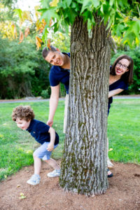 An adult male and female stand behind a tree while a 4 year old runs away from them.