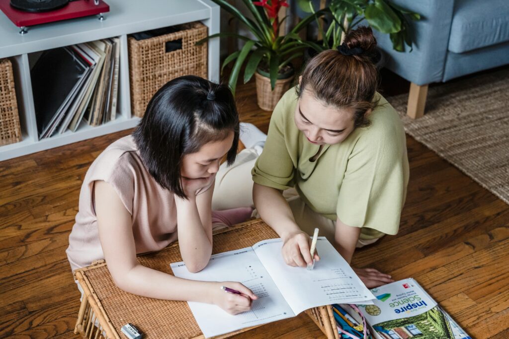 A woman and a girl are lying on the floor working on a homework assignment. They are both focused on a workbook with math problems. Colored pencils and a science textbook are nearby.