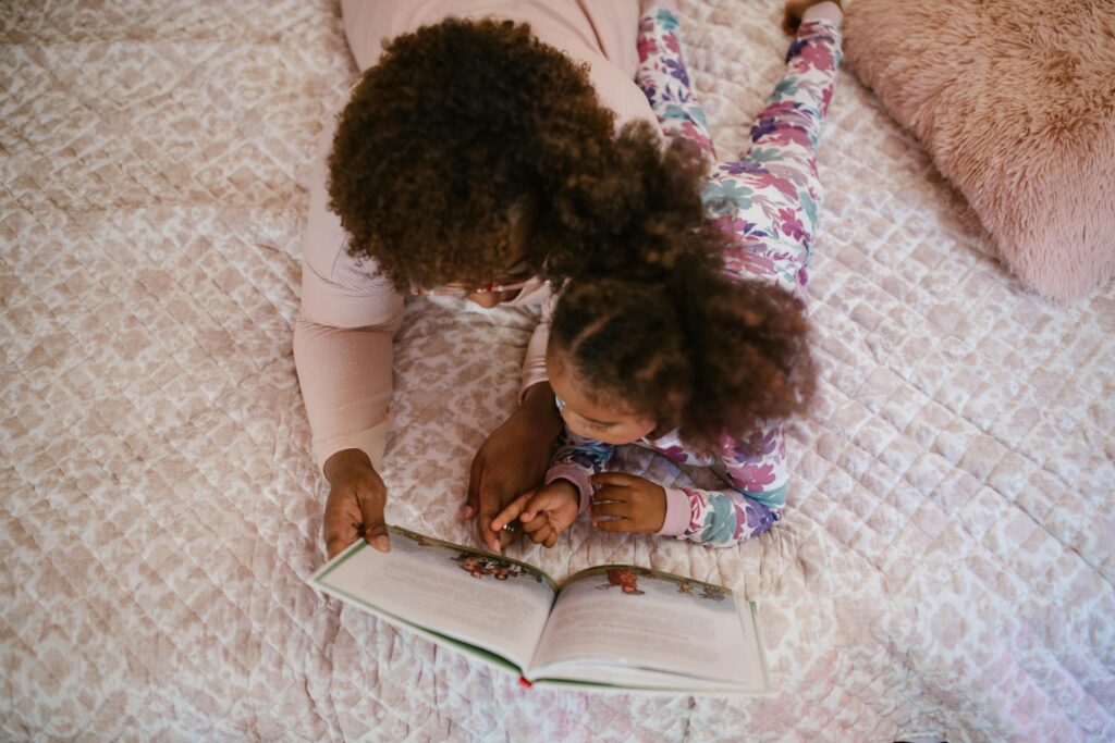 A woman and a young child are lying on a bed together, reading a picture book. The child is pointing at the page while the woman holds the book open.